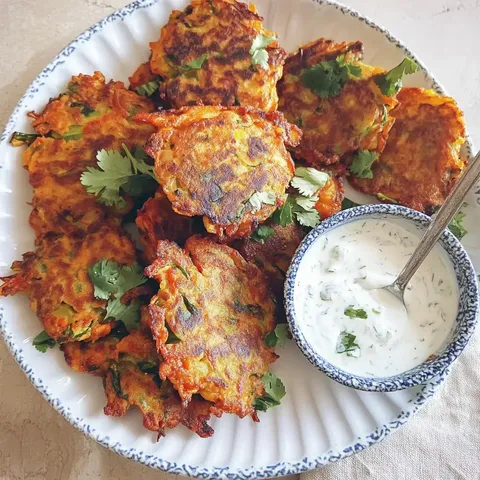 Ein Teller mit knusprigen vegetarischen Pfannkuchen, garniert mit Koriander, neben einer Schüssel mit Joghurtsauce.