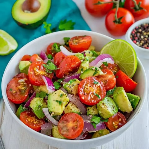 Eine Schüssel frischer Avocado-Tomatensalat mit roten Zwiebeln, Koriander und Limettenscheibe, umgeben von weiteren Tomaten und Avocado.