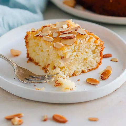 Ein Stück Mandelkuchen auf einem weißen Teller, garniert mit Mandelblättchen, liegt neben einer Gabel.